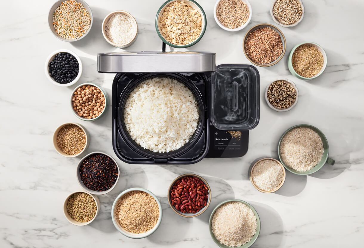 The open rice and grain cooker, with cooked white rice inside. The cooker is surrounded by bowls of various grains, rices and beans.