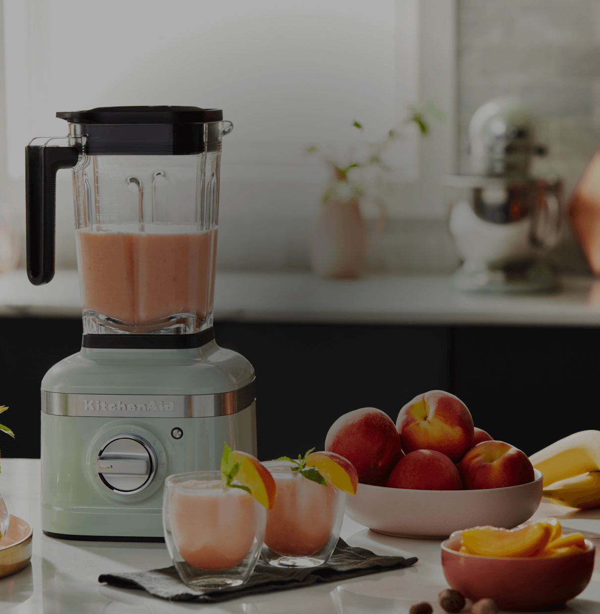 A hand blender, mini food processor and hand mixer on a white countertop with a plate of chocolate chip cookies.