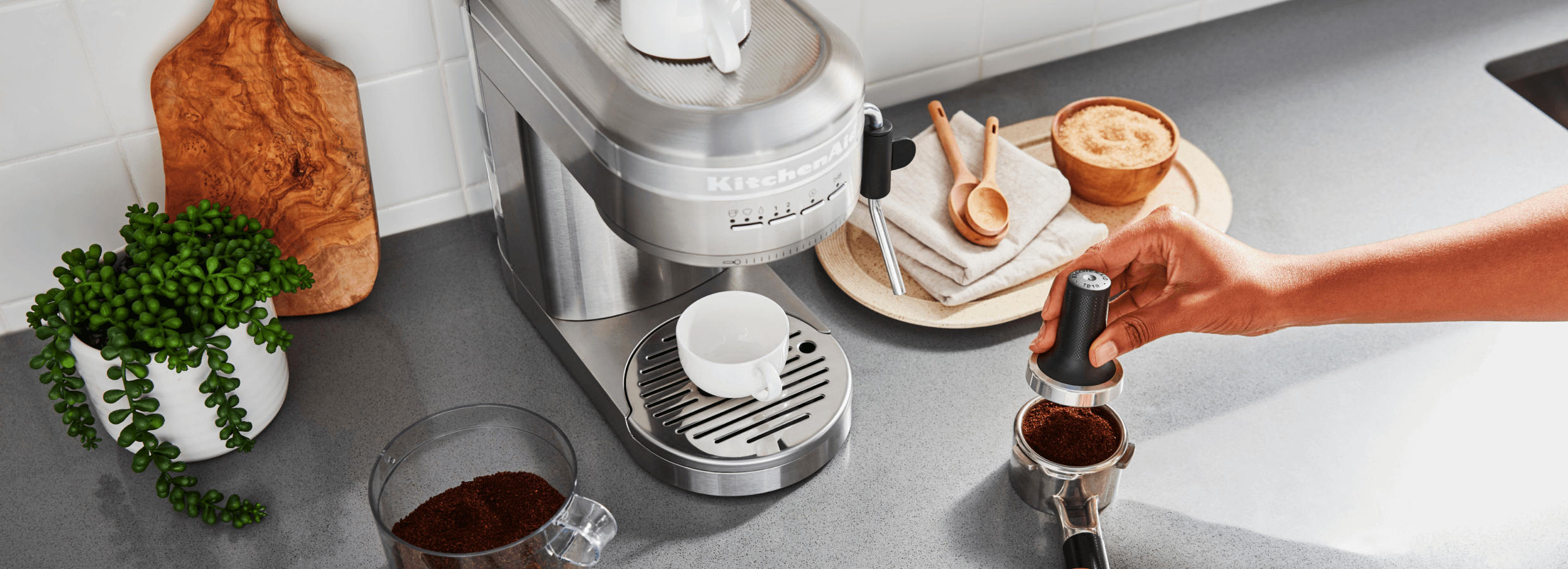 An image of a KitchenAid® Espresso machine, with a person pressing grounds into it.