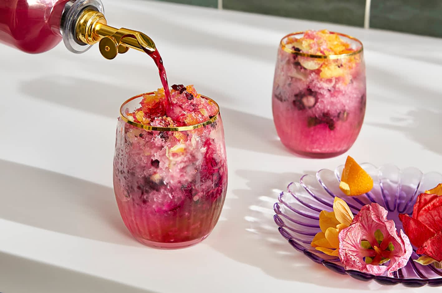 A person pouring hibiscus syrup into a glass filled with shaved ice, a pink beverage and floral garnishes.