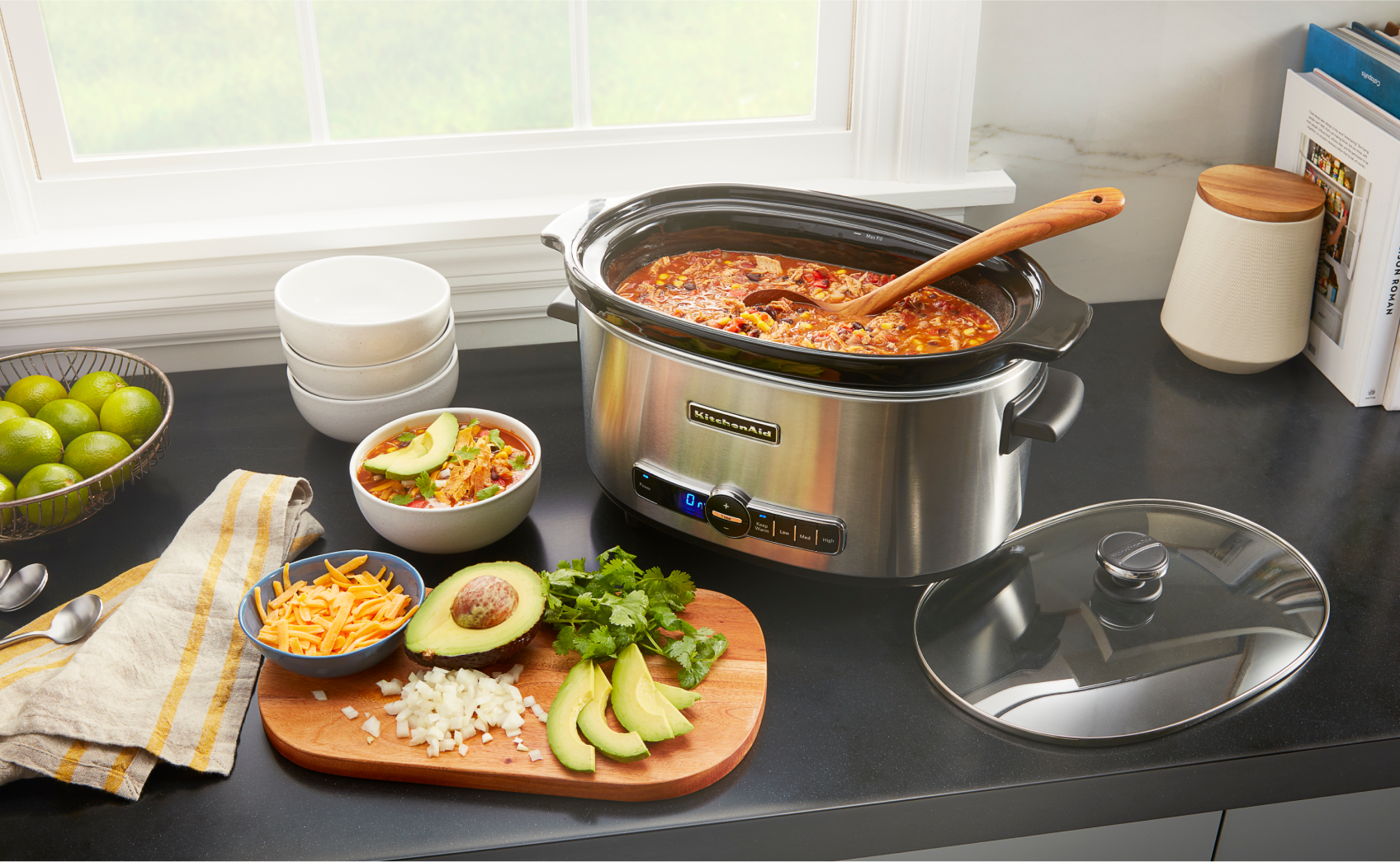 A KitchenAid slow cooker on a countertop, cooking a big batch of chilli. Next to the slow cooker, there are bowls and a cutting board with sliced garnishes like avocado, limes, onion and cheese.