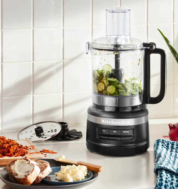 A Matte Black KitchenAid® food processor holding sliced cucumbers on a white countertop.