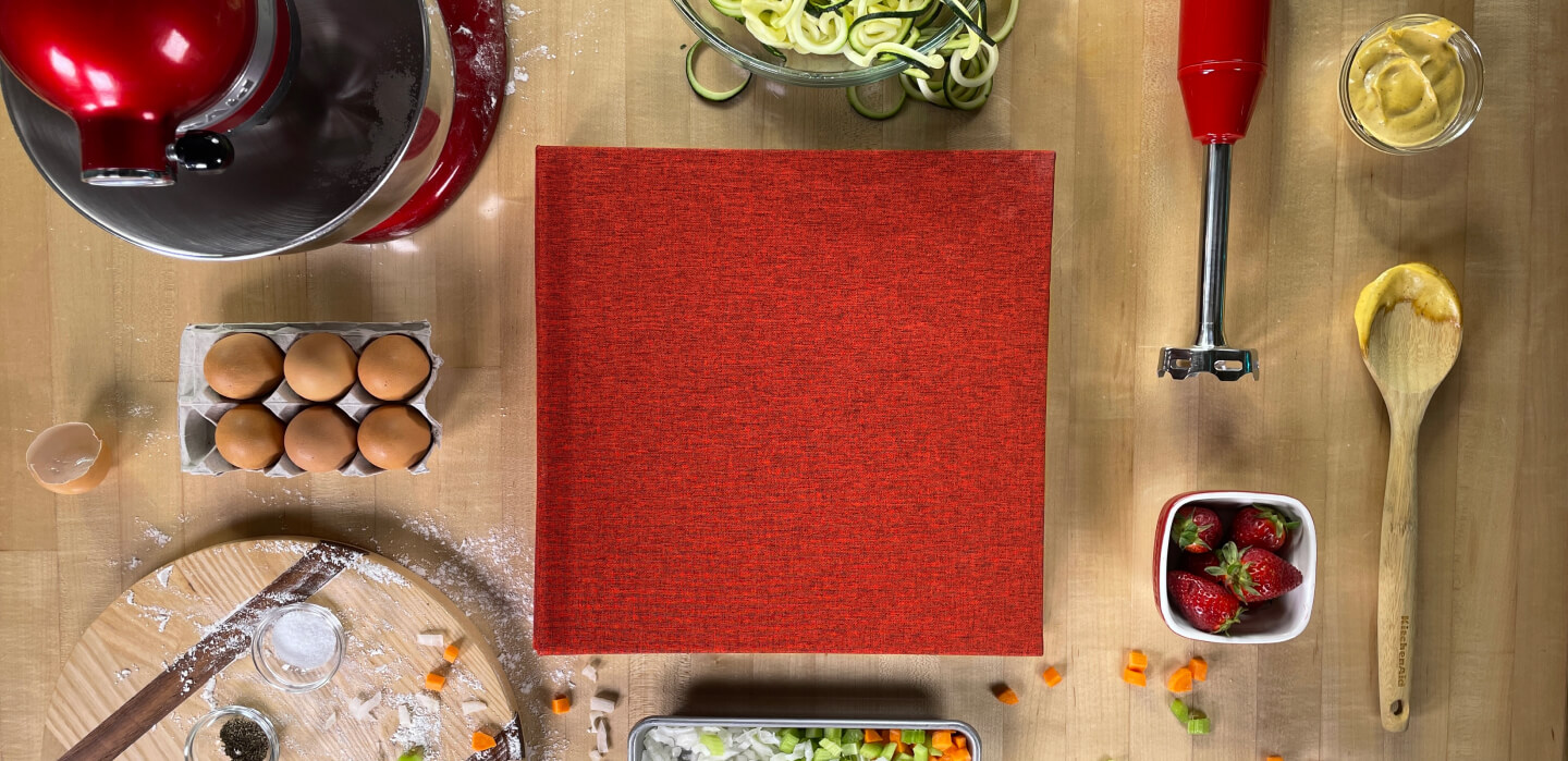 A kitchen countertop scattered with different ingredients and KitchenAid appliances with a red cookbook.