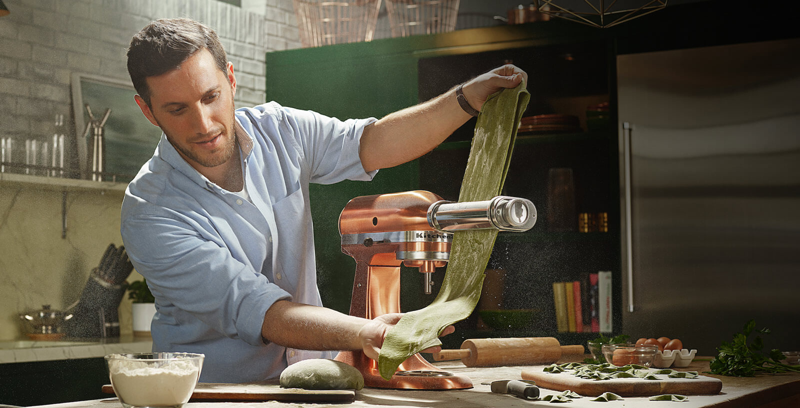 A man running green pasta dough through a pasta sheet roller attached to a Copper Stand Mixer