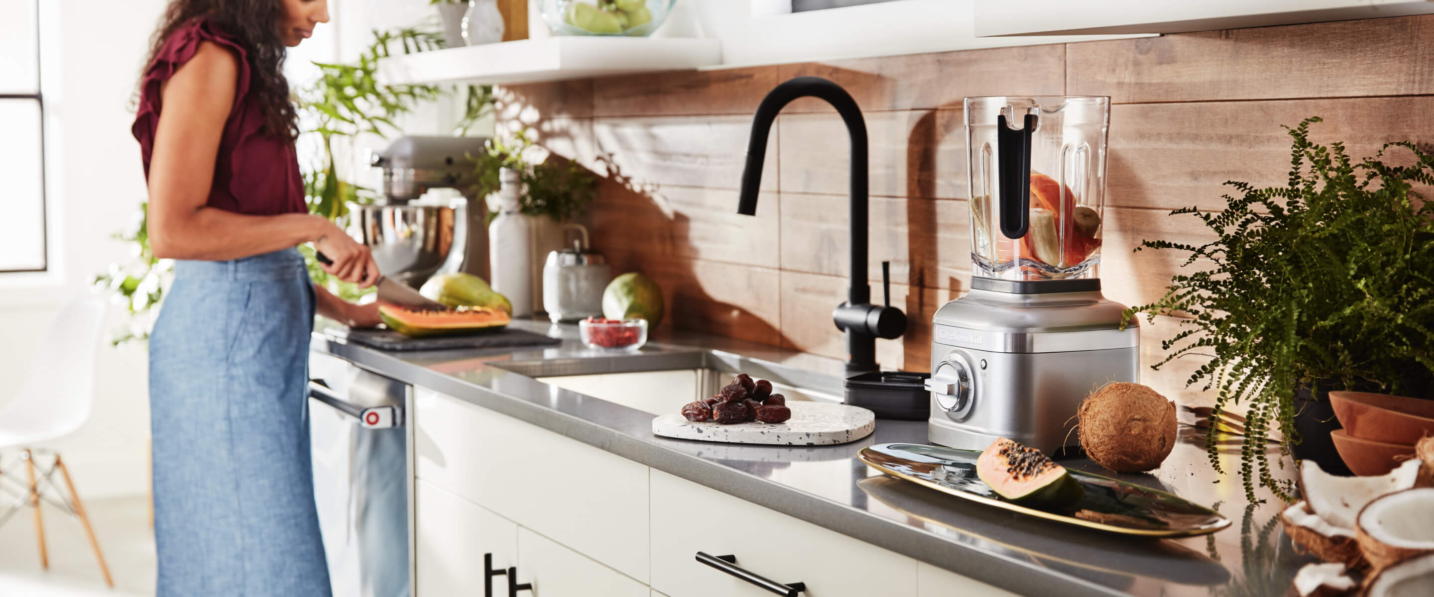Woman using a refurbished KitchenAid® mixer on a countertop with a refurbished KitchenAid® blender nearby.