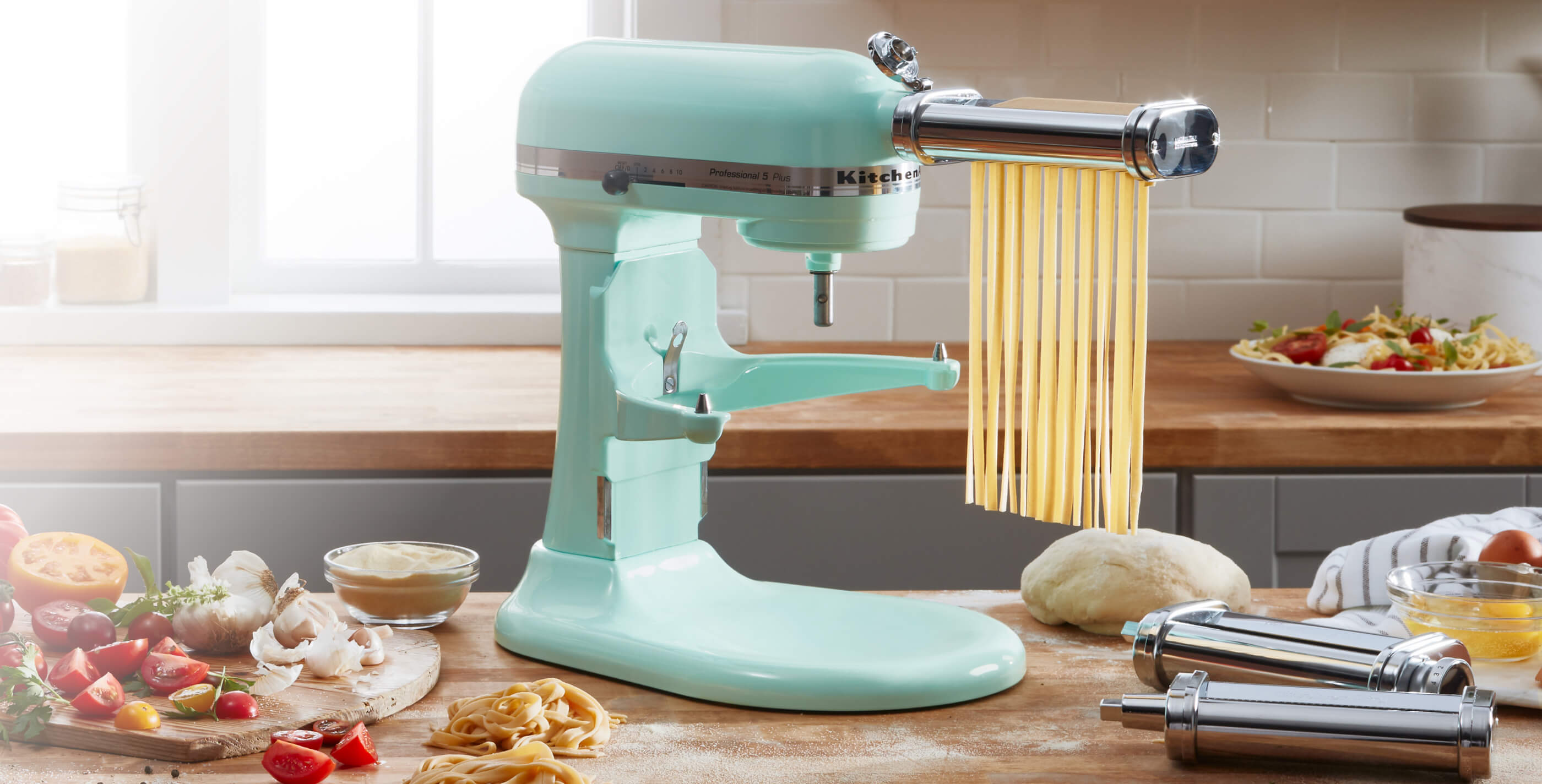 Aqua-colored Stand Mixer with pasta attachment, cutting pasta sheets into linguine on a counter adorned with fresh vegetables.