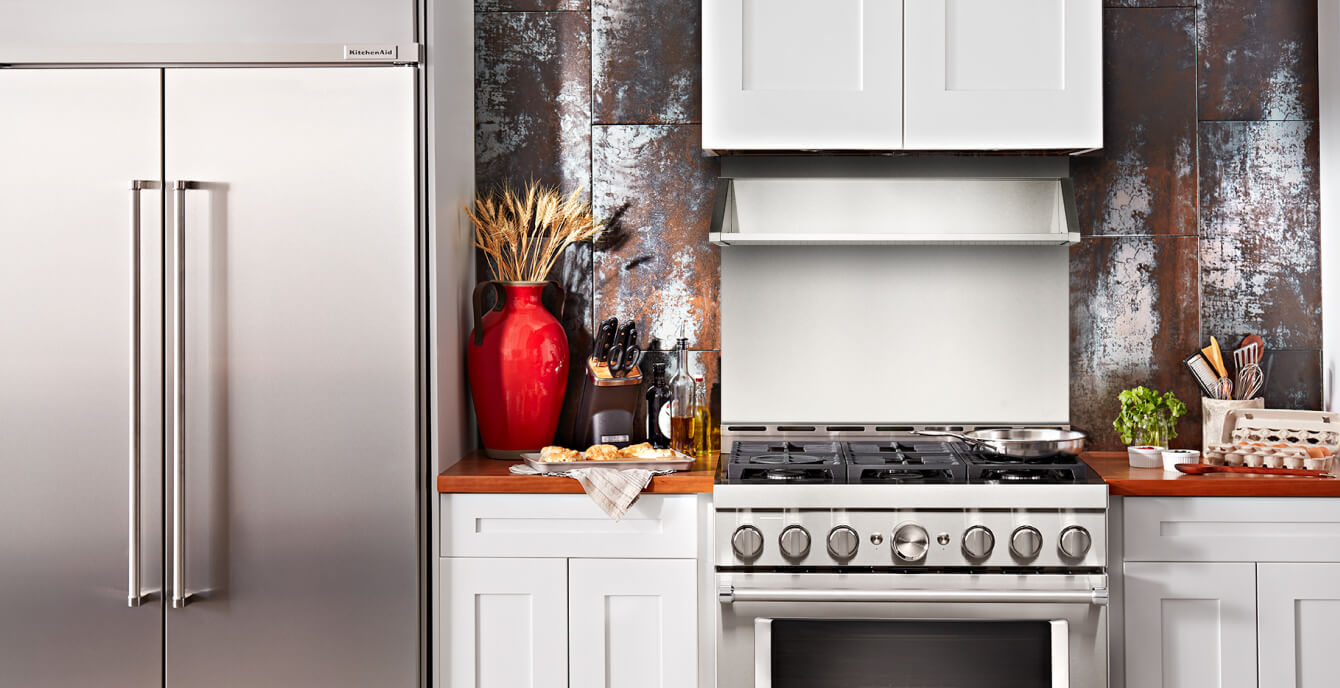 Cropped picture of a kitchen section, with stainless steel refrigerator, commercial-style range and backguard, as well as various cooking items.