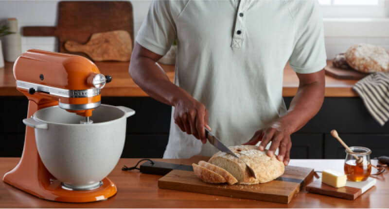 A person slicing a loaf of freshly baked bread, using a KitchenAid® Stand Mixer and the bread baking attachment.
