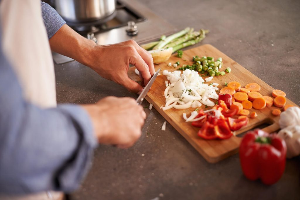 A person dicing white onions on a wooden cutting board along with diced asparagus and tomatoes.