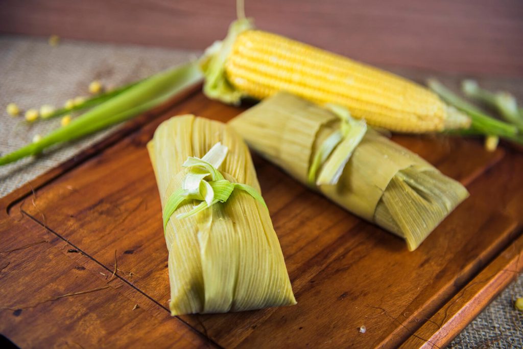 Pamonha resting on a wooden cutting board next to a peeled corn on the cob.