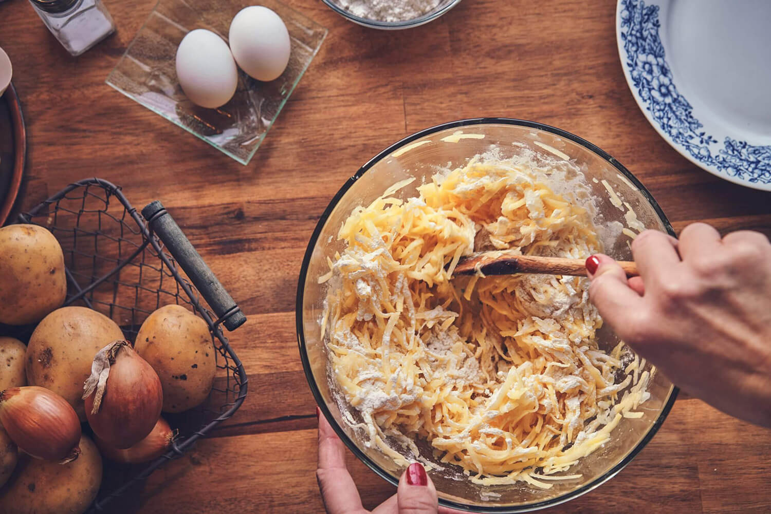 Strong hands stirring shredded potatoes in a glass bowl with a wooden spoon.