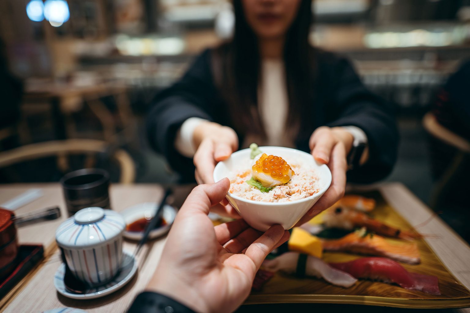 A person handing a small bowl filled with seafood donburi to a young woman.