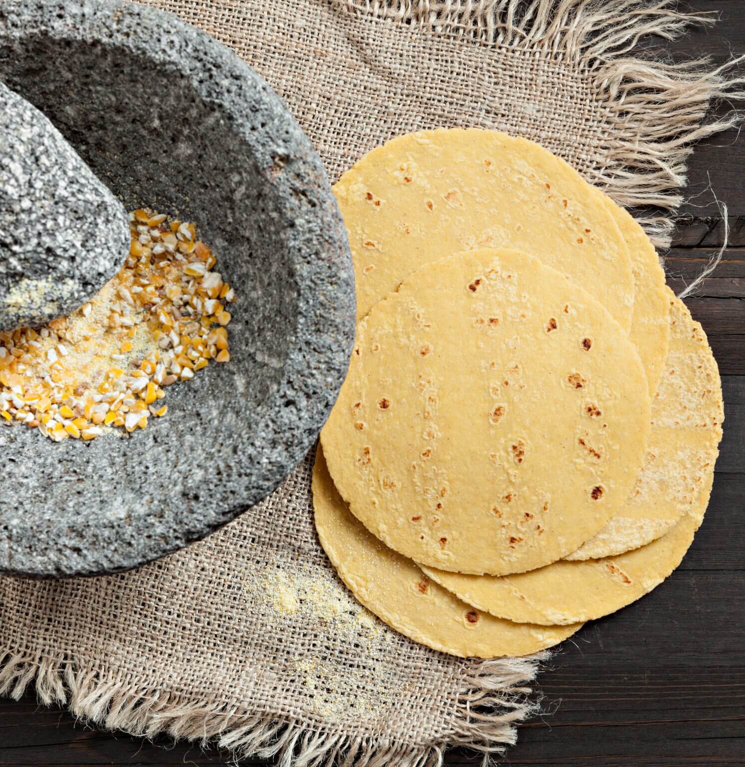 A wooden table with homemade tortillas next to a stone grinder filled with dried corn.