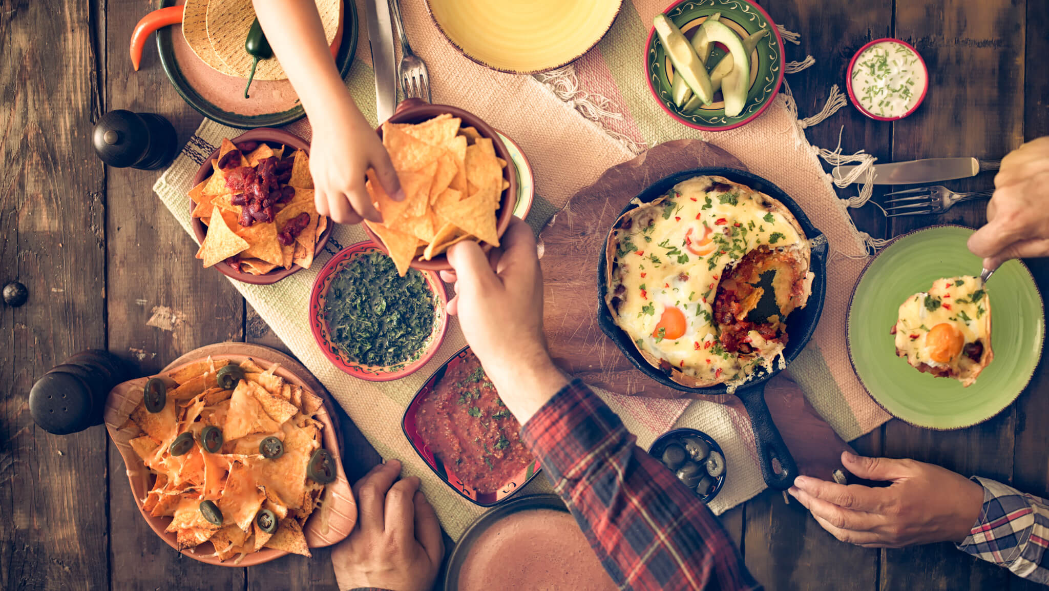 People gathered over a wooden table set with bowls of tortilla chips, dips and salsas.