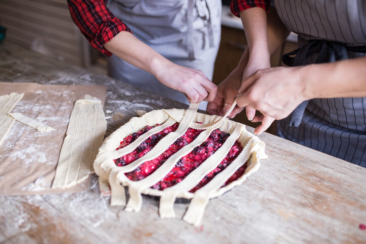 Two people weaving thinly sliced, rolled out dough into an intricate pie crust.