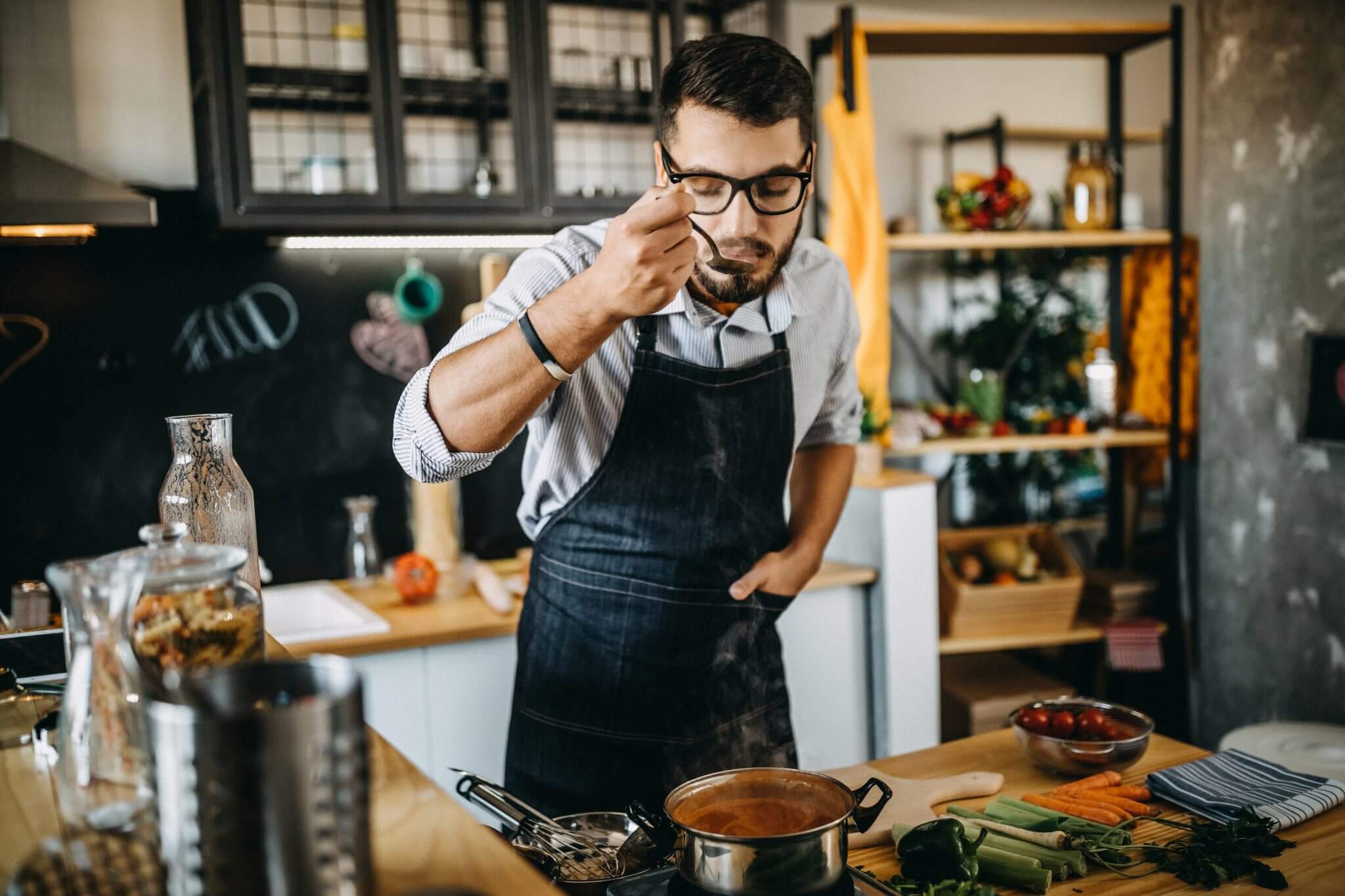 A young man in a denim apron taste-testing his homemade sauce in a pot on the stove.