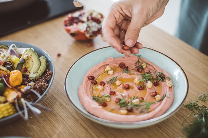 A soft hand sprinkling pomegranate on smooth hummus with herbs and whole chickpeas.