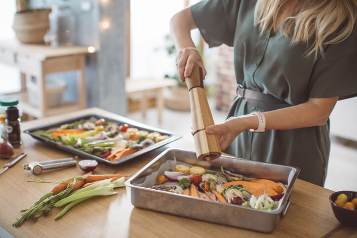 A woman seasoning chopped vegetables in a baking tray with a wooden pepper grinder.