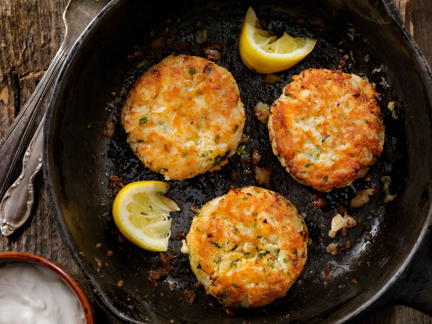 Three crab cakes frying in a skillet with sliced lemon.