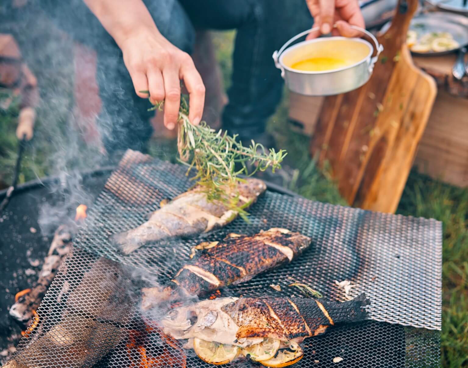 A person grilling three freshly caught fish stuffed with sliced lemons over an open fire pit.
