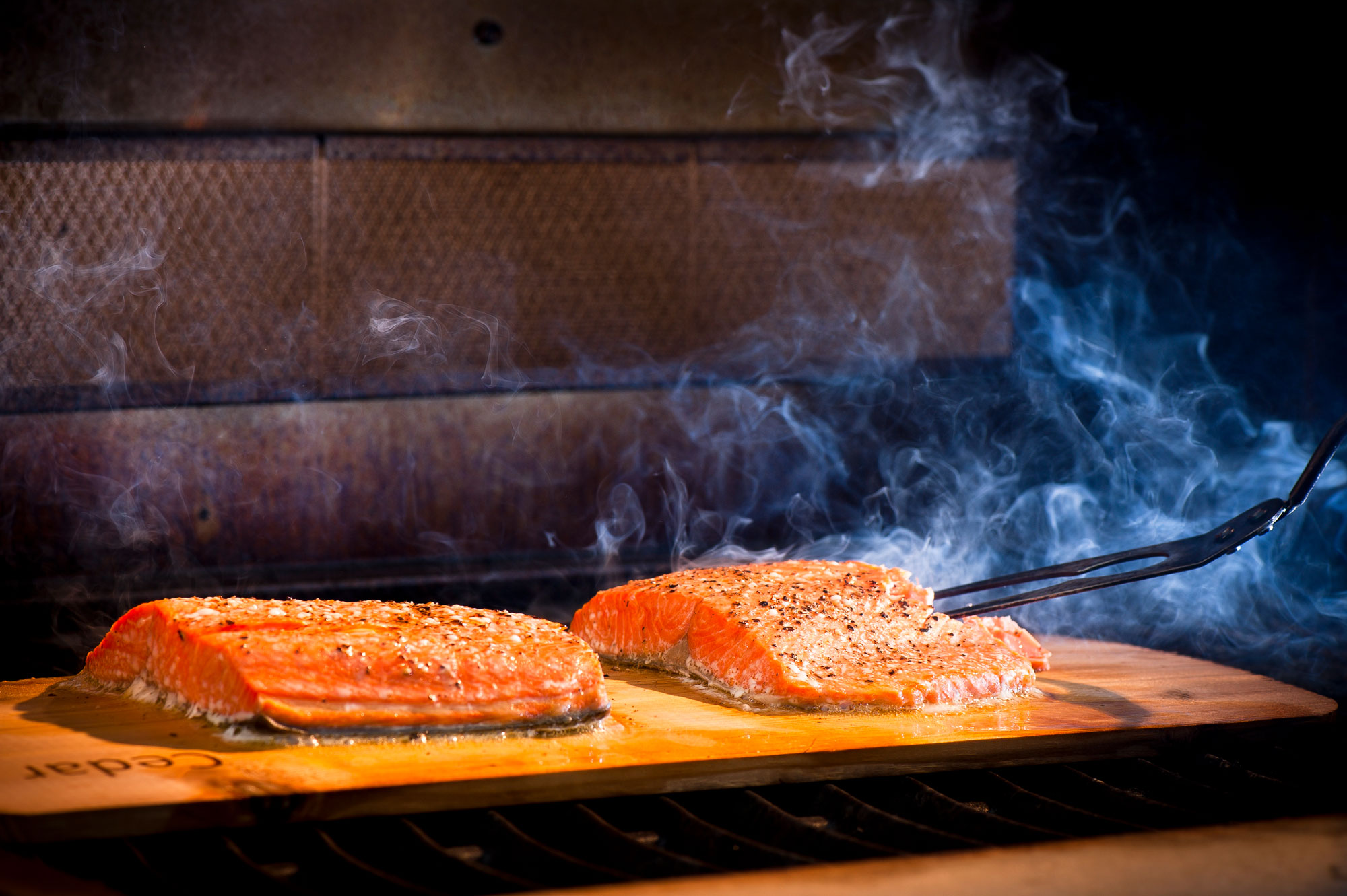 Two salmon fillets cooking on a thin, wooden plank.