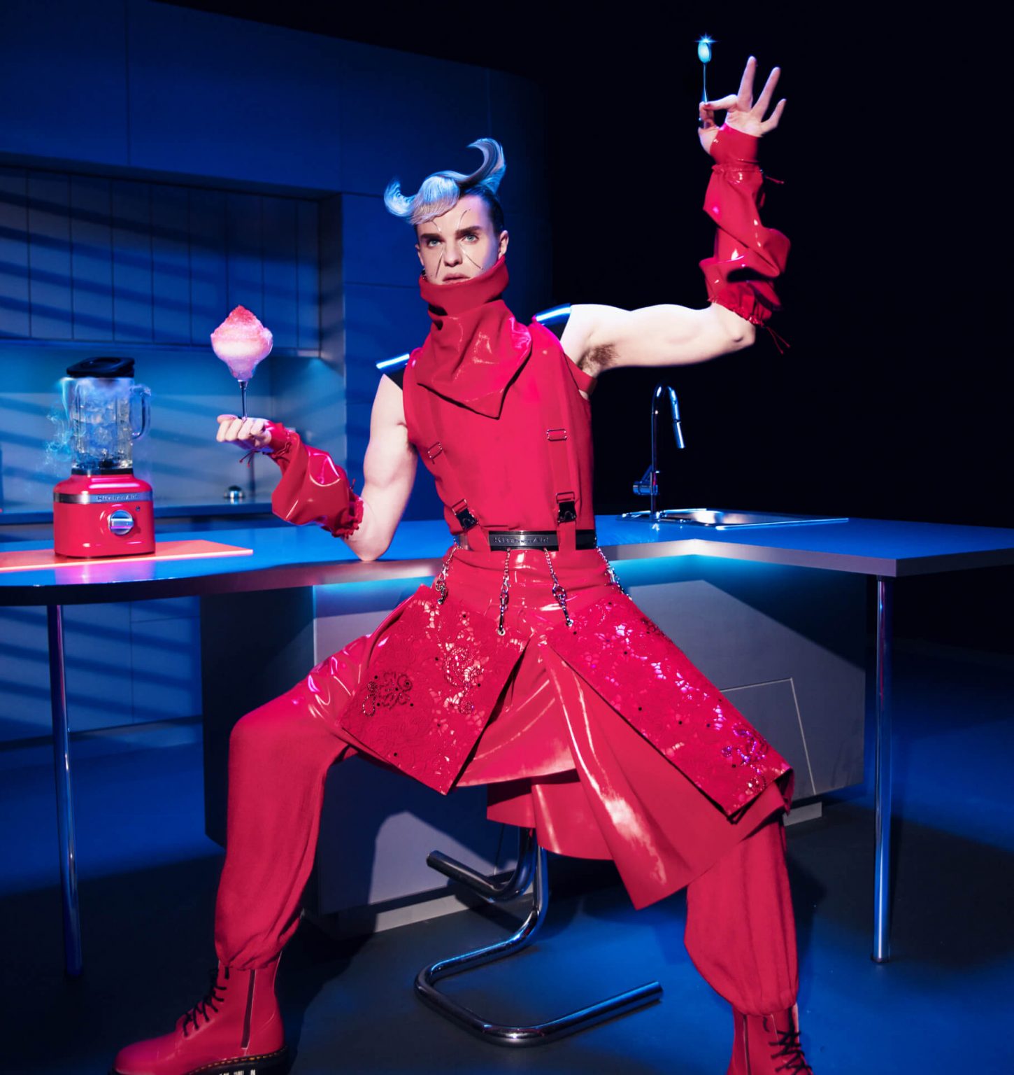 A model posing in a kitchen wearing Look Five while holding a glass filled with whipped pink cream near the K400 Blender in Hibiscus.