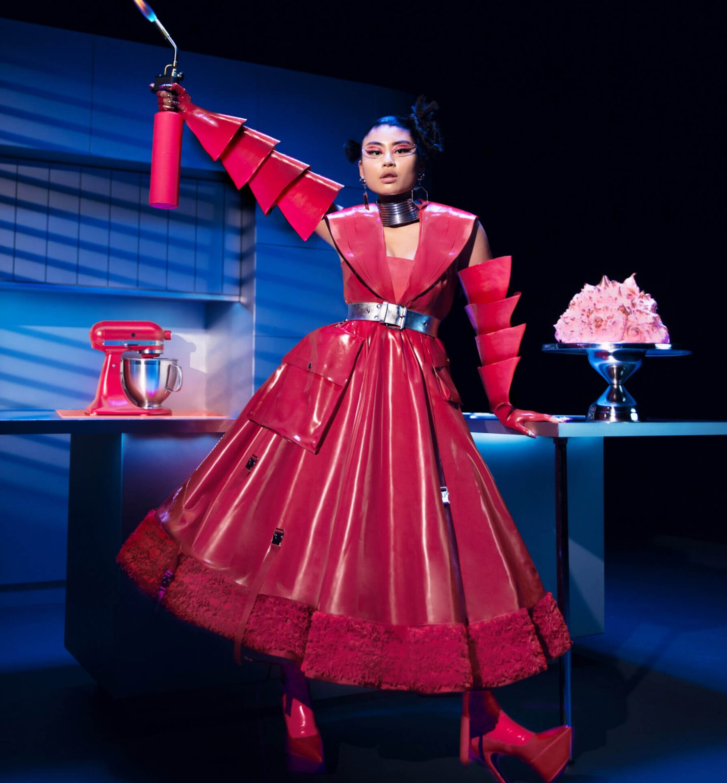 A model posing in a kitchen wearing Look Seven while holding a lit blow torch near a toasted meringue dessert.