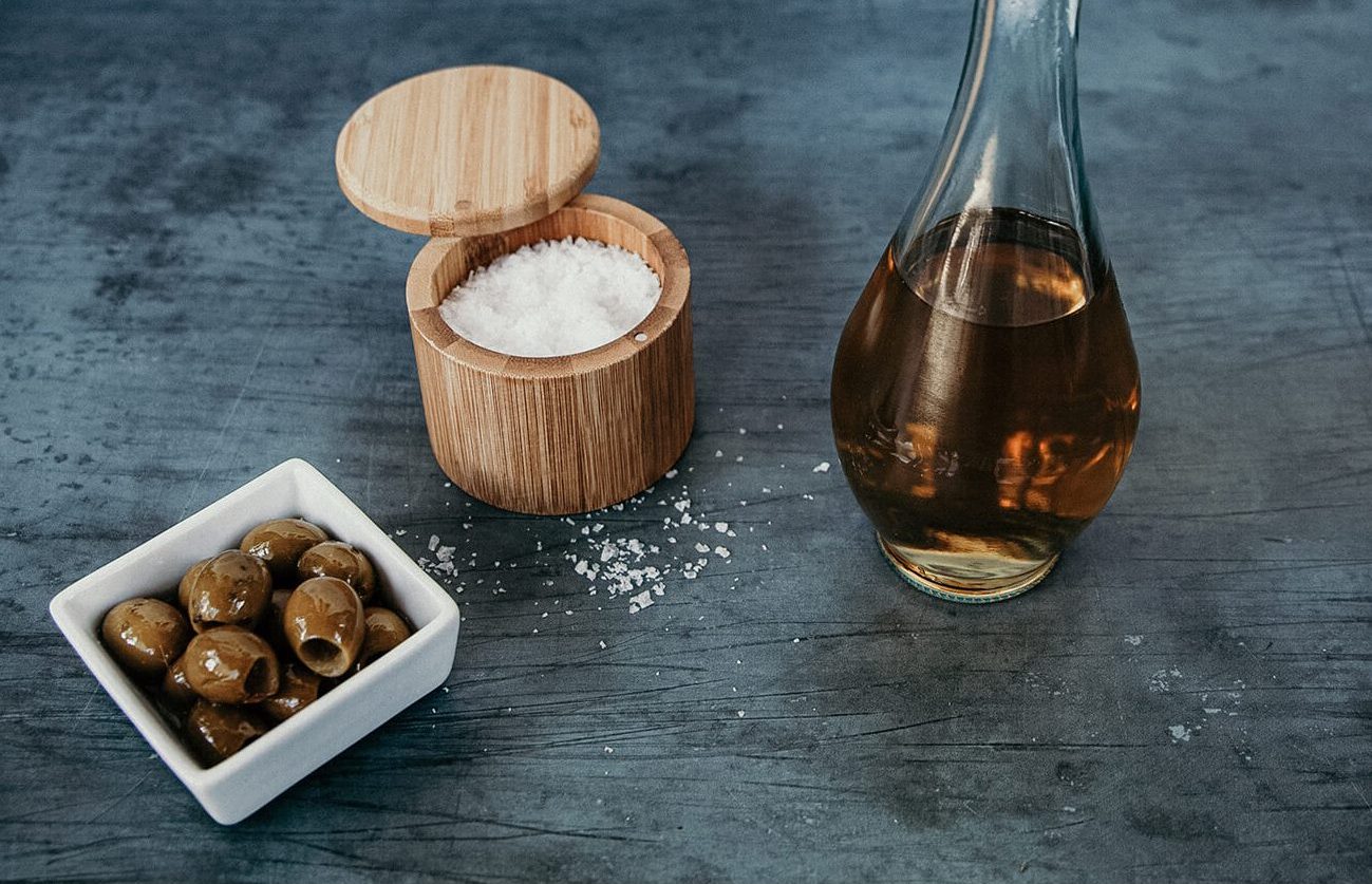 A bottle of vinegar, a dish of olives and a container of kosher salt resting on a table.