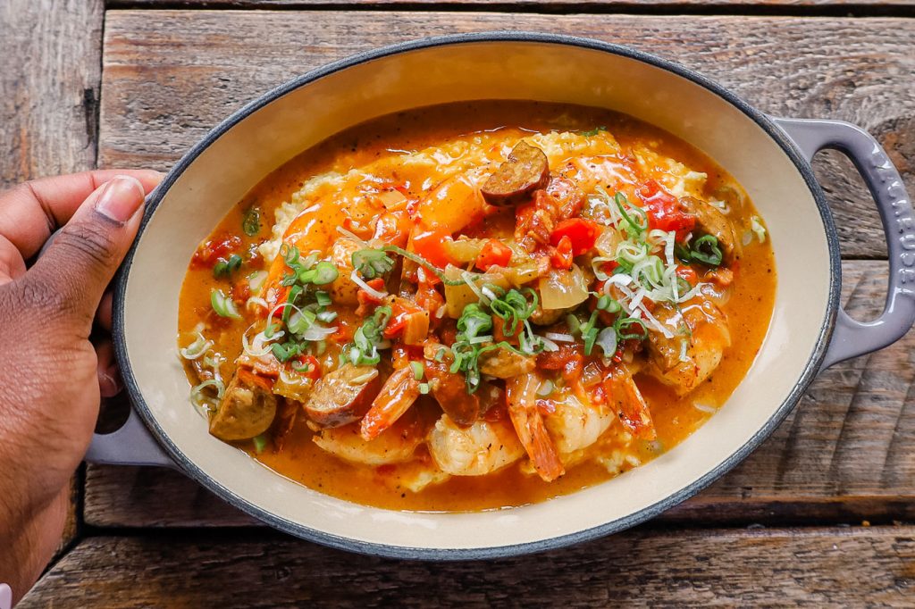 A person holding an oval dish filled with steaming cajun shrimp and grits.