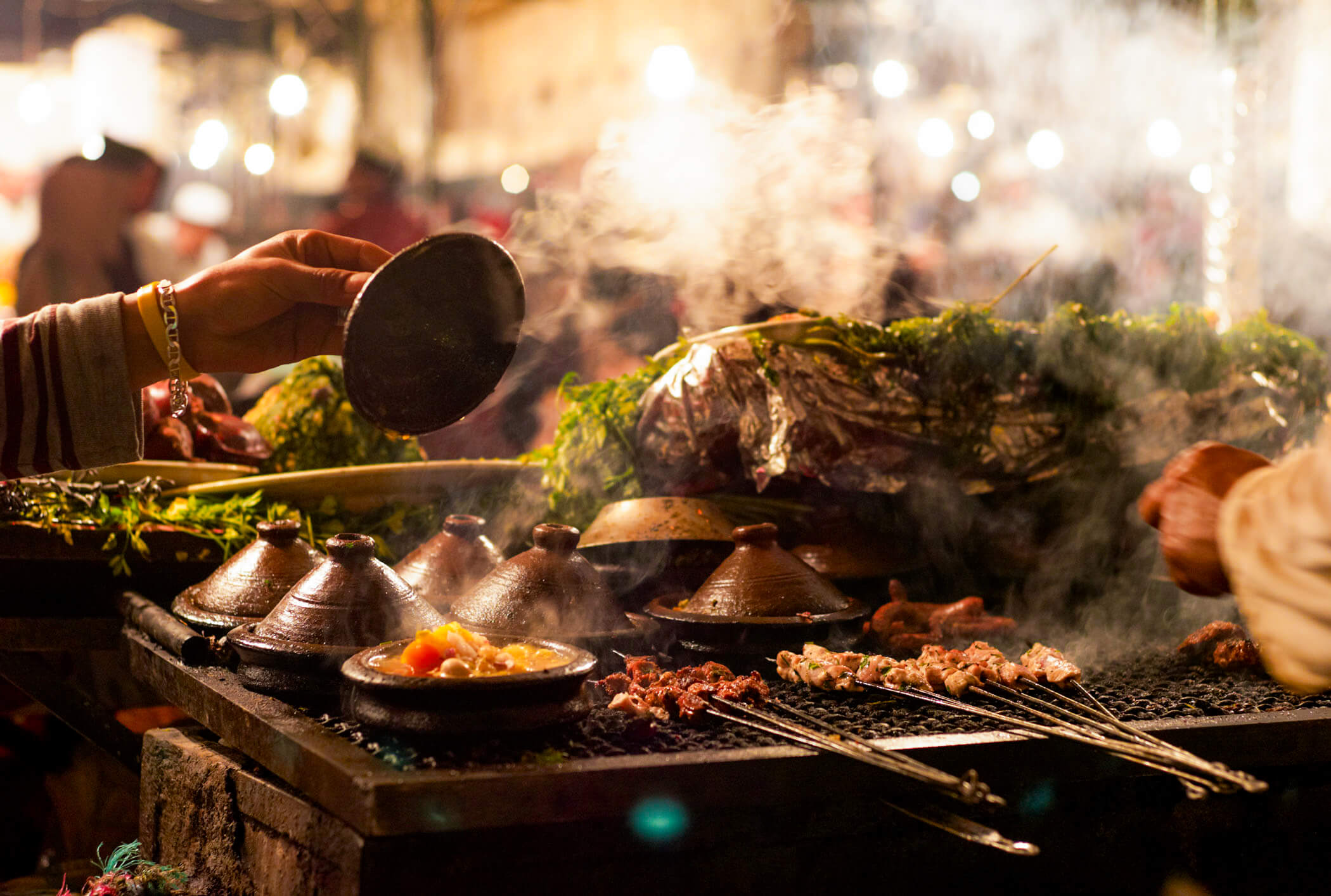 A woman checking her tagine pots as they sit on a large grill with various meats and kebabs.
