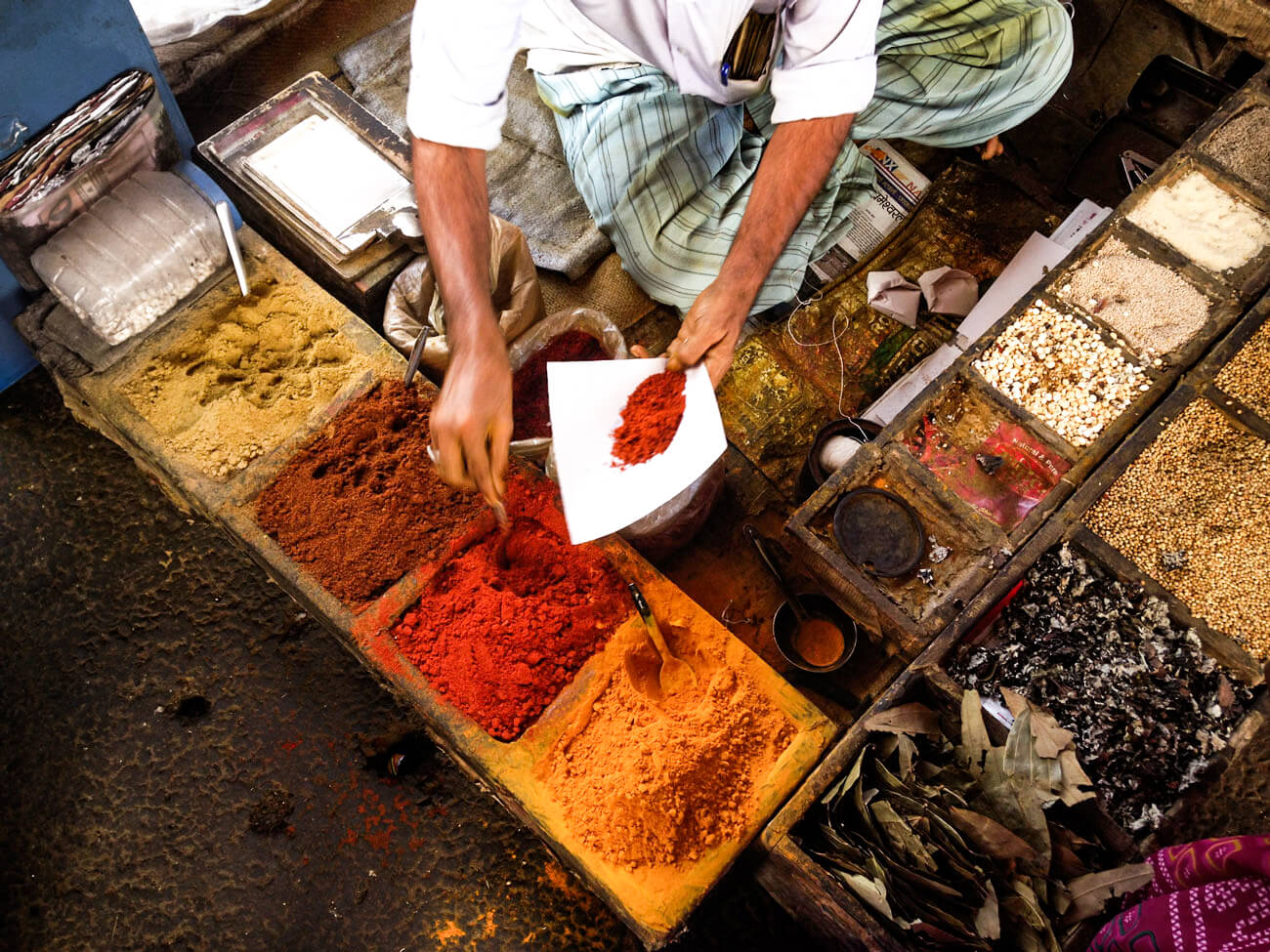 A person collecting a red spice amongst a variety of colorful spices, seasonings and dried herbs.