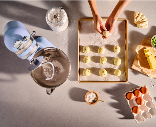 A person rolling cookie dough into balls and placing them on a cookie sheet next to a Blue Salt stand mixer.