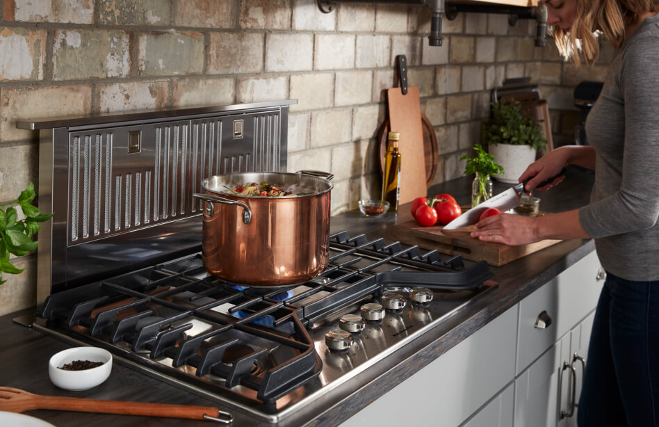 Bronze pot resting on center grate of a cooktop and person slicing tomatoes on a cutting board Bronze pot resting on center grate of a cooktop and person slicing tomatoes on a cutting board