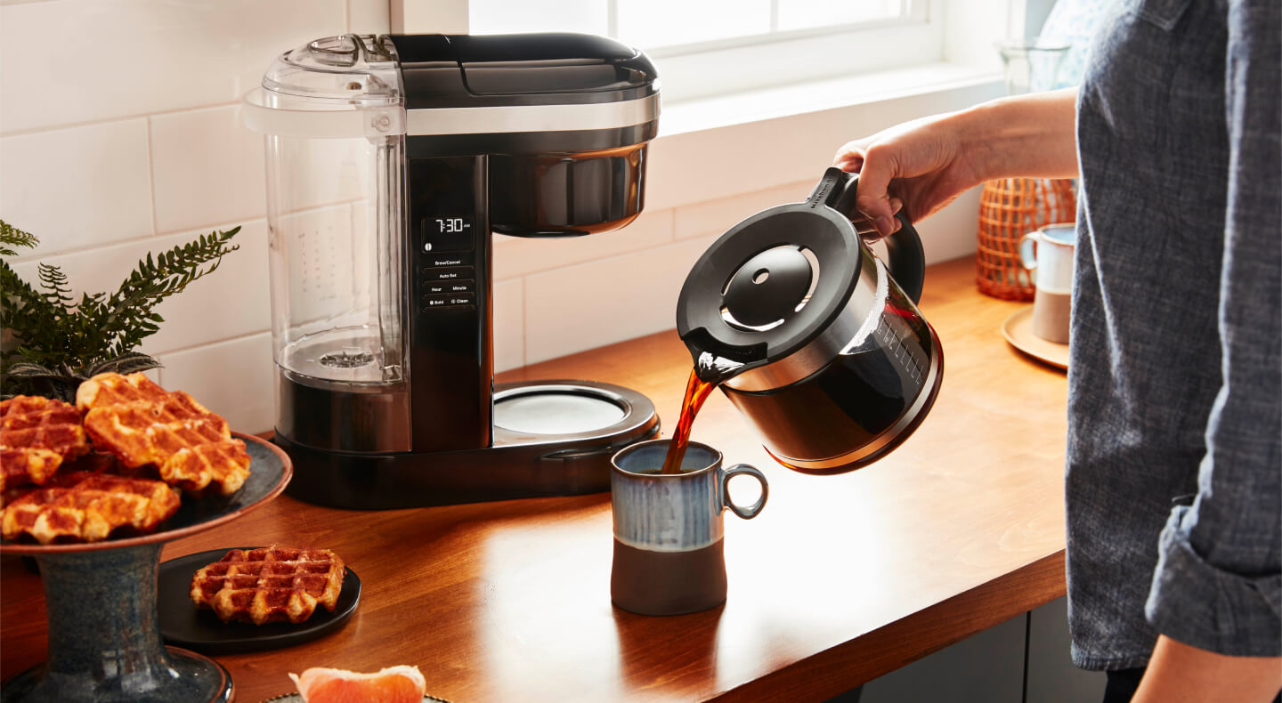 Person pouring coffee from a KitchenAid® coffee pot into a ceramic mug