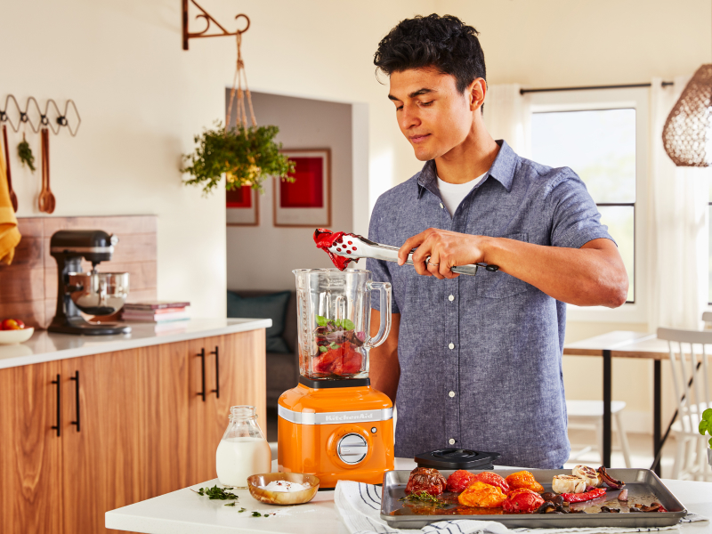 A man adding roasted vegetables to a KitchenAid® blender in a modern kitchen. A man adding roasted vegetables to a KitchenAid® blender in a modern kitchen.