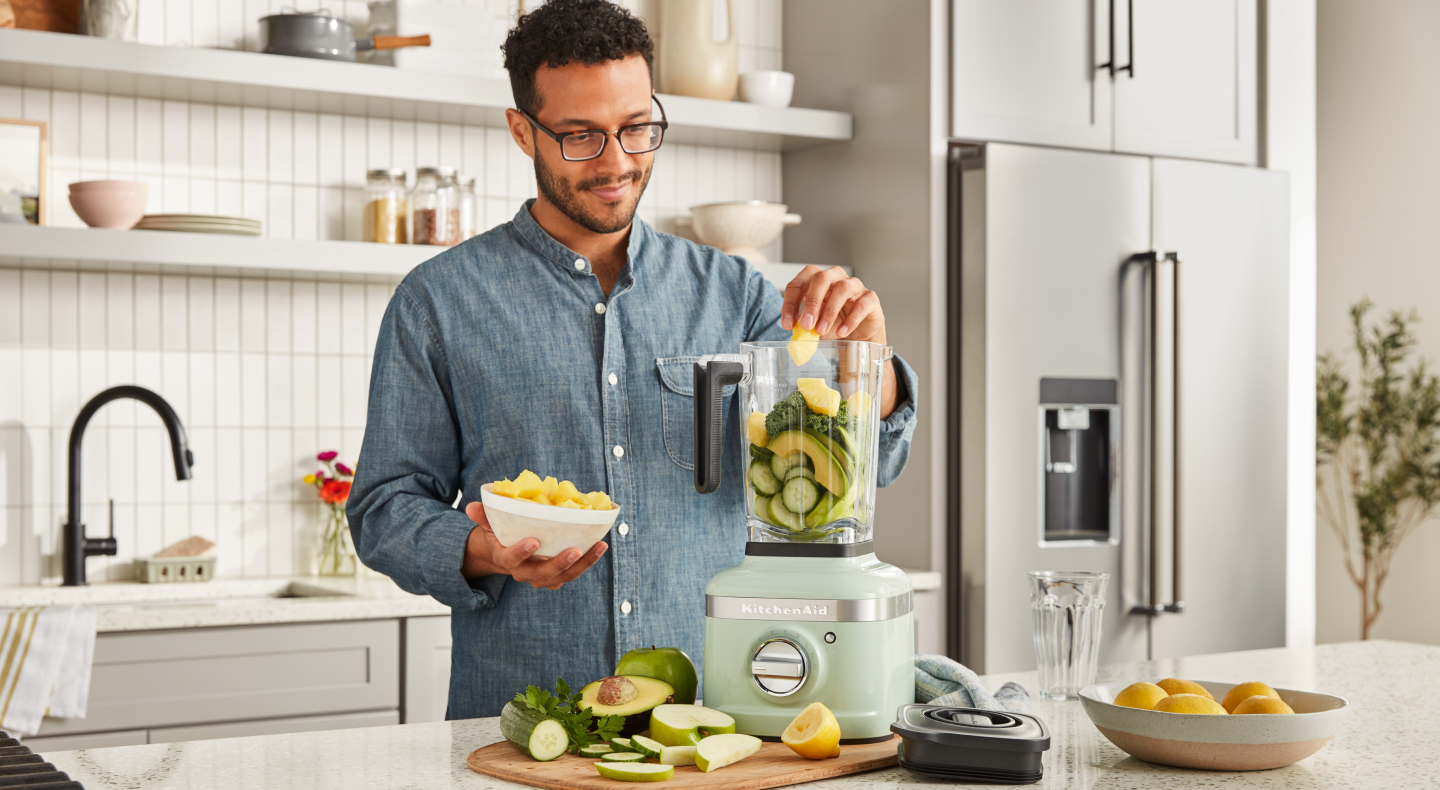 A man putting fresh vegetables into a KitchenAid® blender in a modern kitchen. A man putting fresh vegetables into a KitchenAid® blender in a modern kitchen.