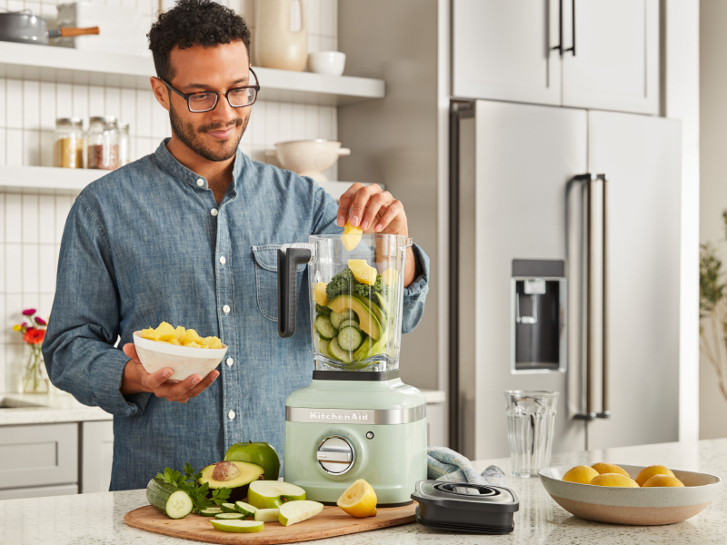 A man putting fresh vegetables into a KitchenAid® blender in a modern kitchen. A man putting fresh vegetables into a KitchenAid® blender in a modern kitchen.