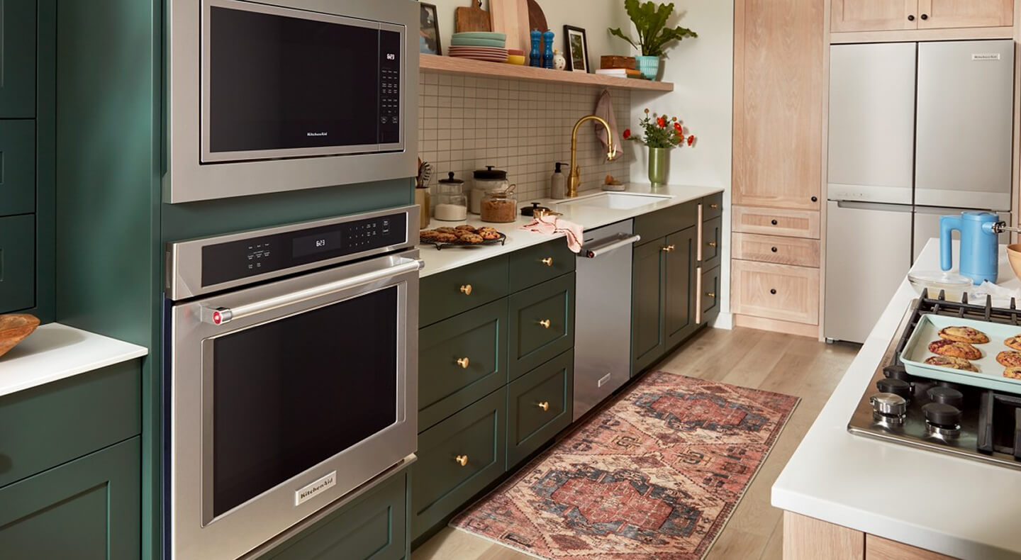 A galley kitchen with forest green cabinetry, white countertops, stainless steel appliances and white tile backsplash on one side, and a kitchen island with white countertops and a stove top on the other A galley kitchen with forest green cabinetry, white countertops, stainless steel appliances and white tile backsplash on one side, and a kitchen island with white countertops and a stove top on the other