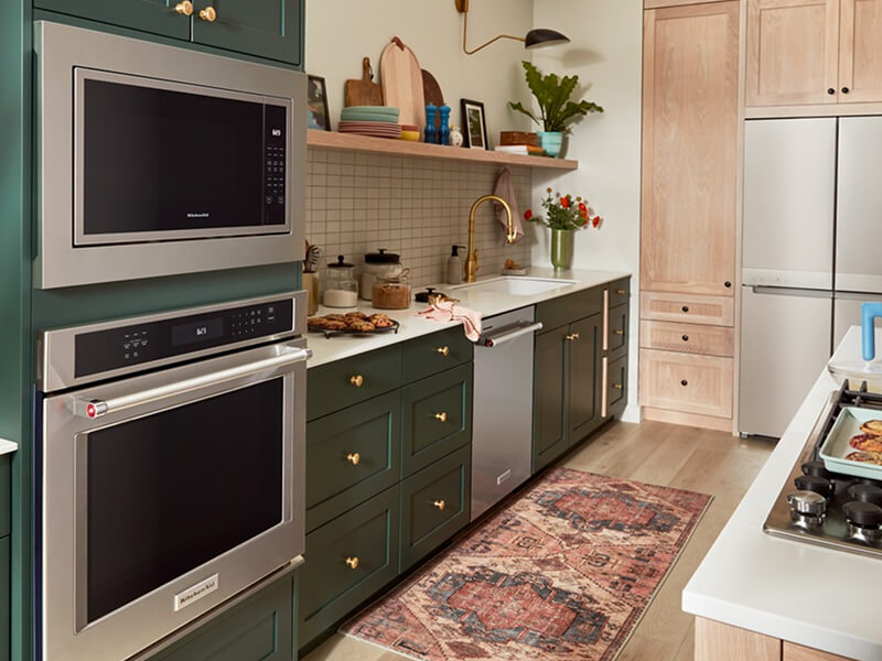 A galley kitchen with forest green cabinetry, white countertops, stainless steel appliances and white tile backsplash on one side, and a kitchen island with white countertops and a stove top on the other A galley kitchen with forest green cabinetry, white countertops, stainless steel appliances and white tile backsplash on one side, and a kitchen island with white countertops and a stove top on the other