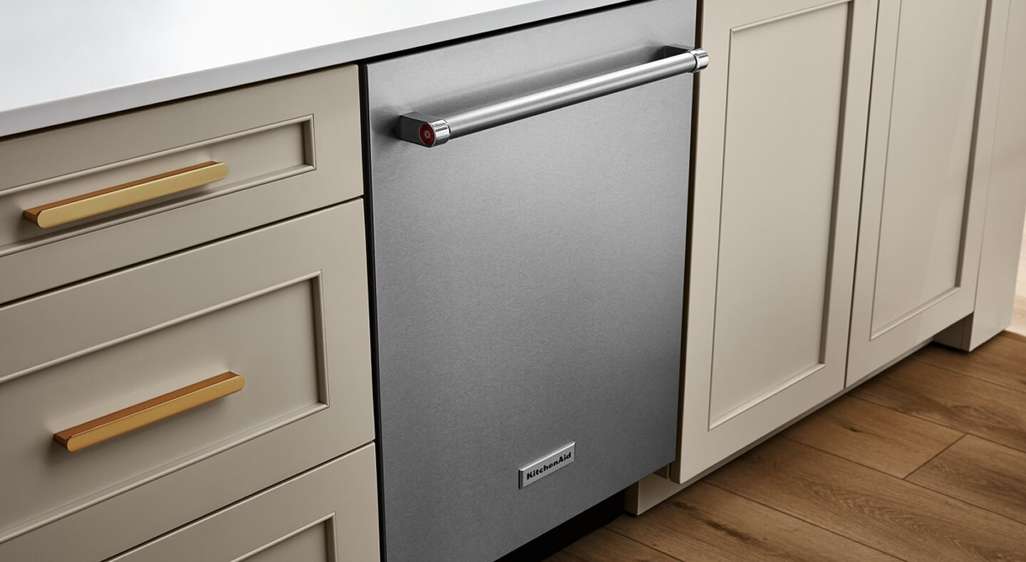 A stainless steel top control dishwasher that’s built into tan cabinetry with gold handles and white countertops
