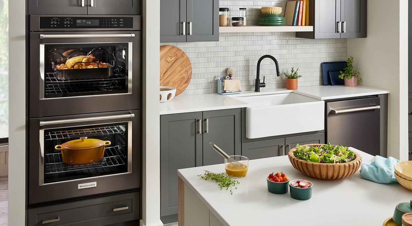 A kitchen with a double oven, apron sink, gray cabinets and a dishwasher against one wall, and an island with a white countertop across the way A kitchen with a double oven, apron sink, gray cabinets and a dishwasher against one wall, and an island with a white countertop across the way