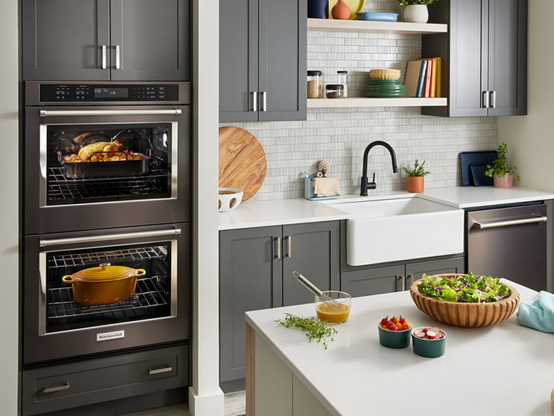 A kitchen with a double oven, apron sink, gray cabinets and a dishwasher against one wall, and an island with a white countertop across the way A kitchen with a double oven, apron sink, gray cabinets and a dishwasher against one wall, and an island with a white countertop across the way