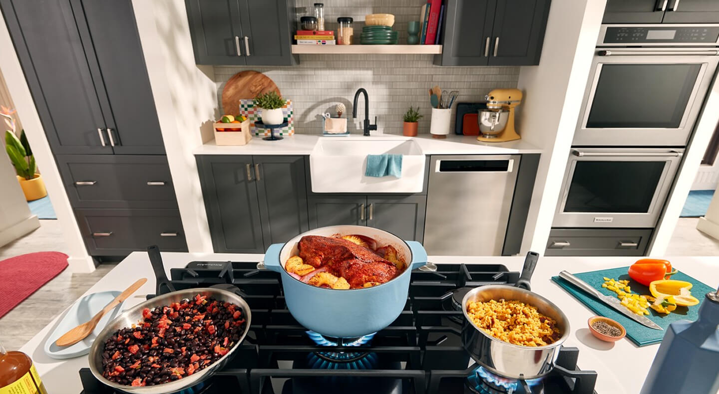 A galley kitchen with dark gray cabinets, dishwasher and double oven on one side, and an island with a stovetop loaded with full pots and pans on the other A galley kitchen with dark gray cabinets, dishwasher and double oven on one side, and an island with a stovetop loaded with full pots and pans on the other