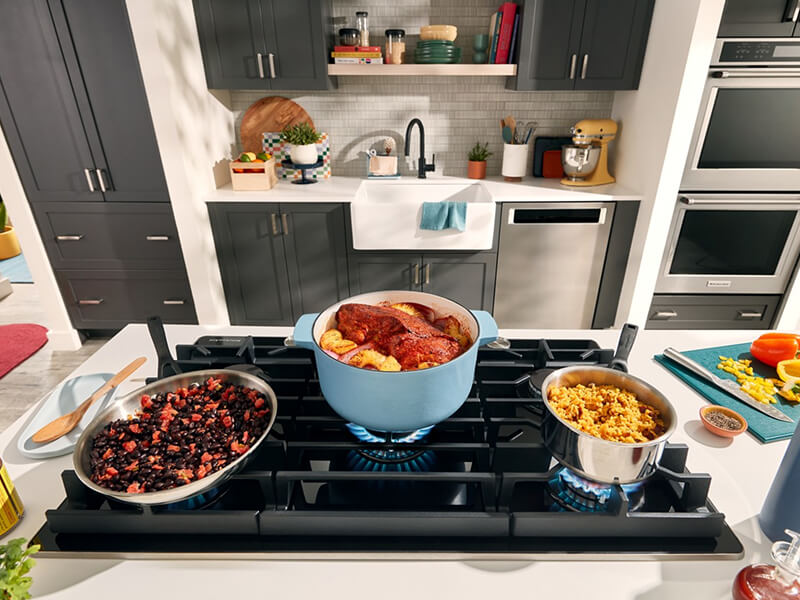 A galley kitchen with dark gray cabinets, dishwasher and double oven on one side, and an island with a stovetop loaded with full pots and pans on the other A galley kitchen with dark gray cabinets, dishwasher and double oven on one side, and an island with a stovetop loaded with full pots and pans on the other