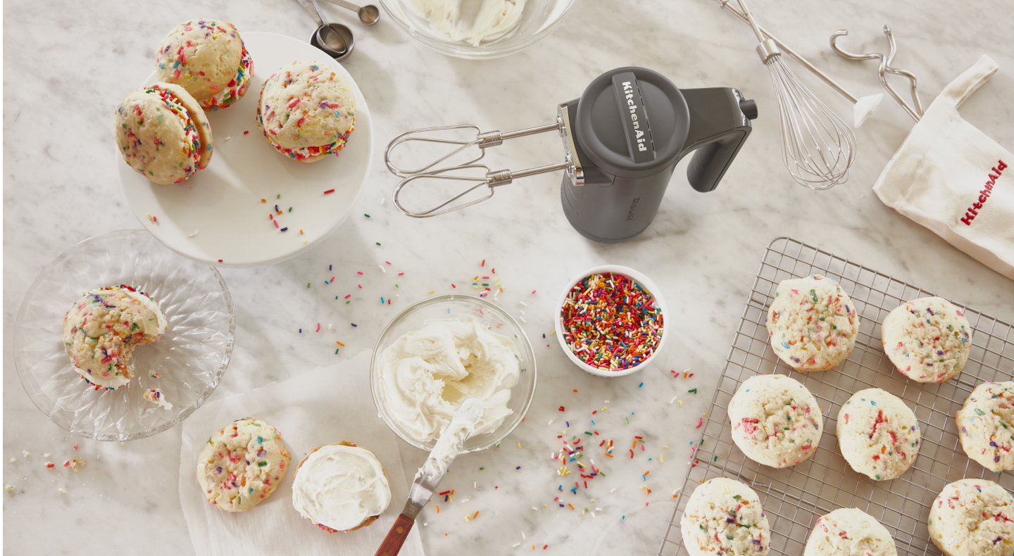 Cordless KitchenAid® hand mixer next to creamy frosting and fresh baked sandwich cookies with colorful sprinkles. Cordless KitchenAid® hand mixer next to creamy frosting and fresh baked sandwich cookies with colorful sprinkles.