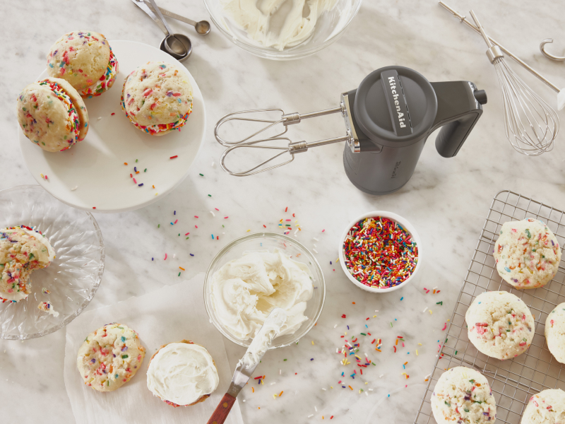 Cordless KitchenAid® hand mixer next to creamy frosting and fresh baked sandwich cookies with colorful sprinkles. Cordless KitchenAid® hand mixer next to creamy frosting and fresh baked sandwich cookies with colorful sprinkles.