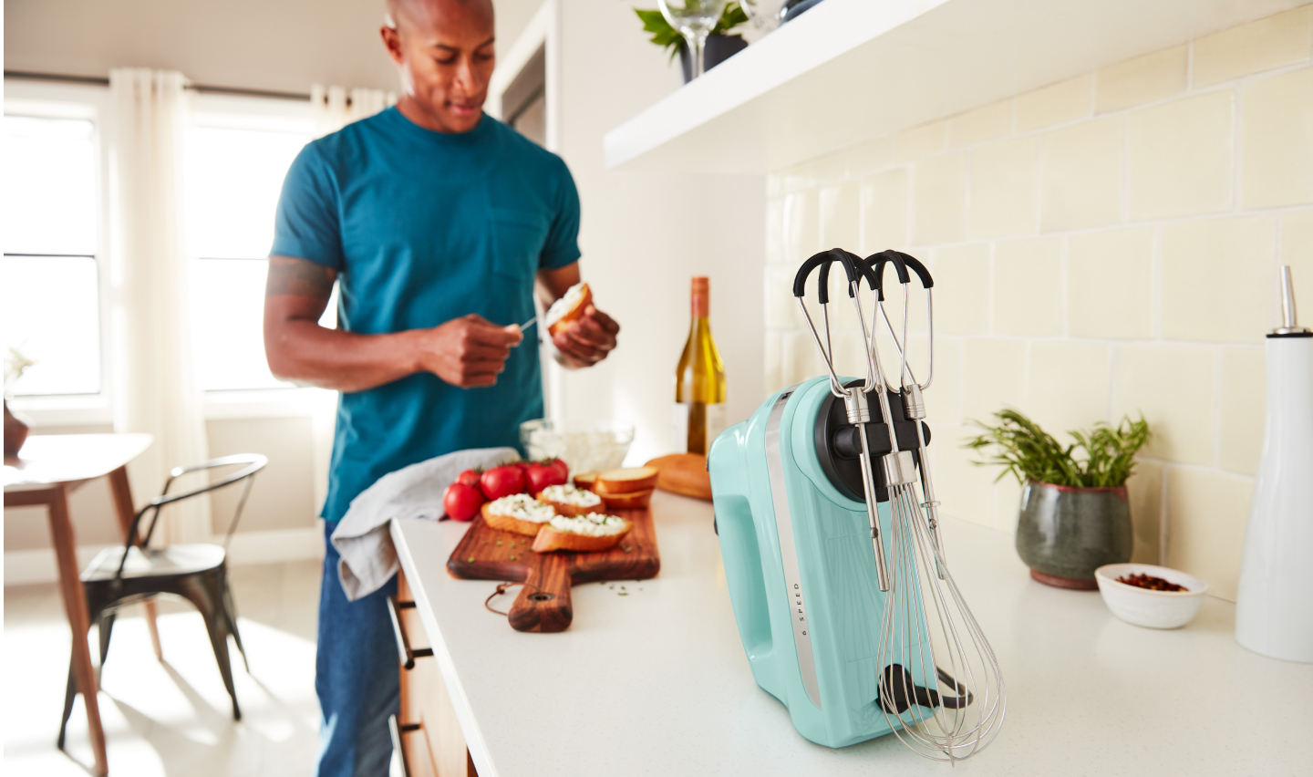 A man in a modern kitchen spreading goat cheese on a crostini next to a KitchenAid® hand mixer.  A man in a modern kitchen spreading goat cheese on a crostini next to a KitchenAid® hand mixer.