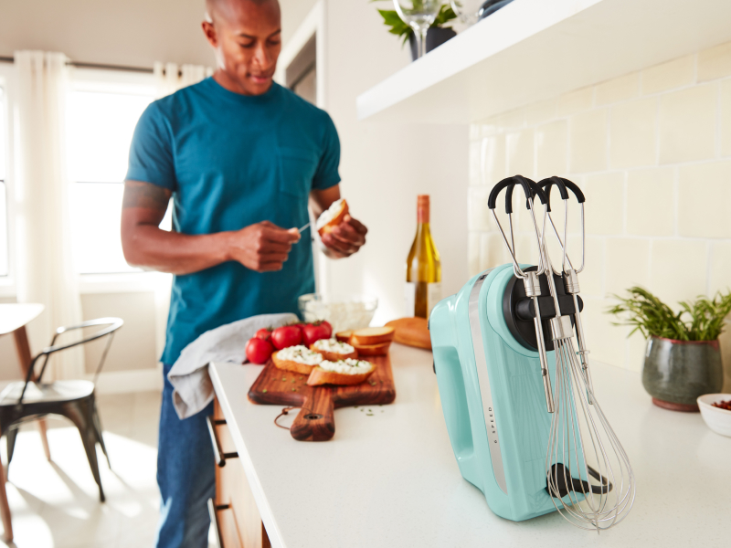 A man in a modern kitchen spreading goat cheese on a crostini next to a KitchenAid® hand mixer.  A man in a modern kitchen spreading goat cheese on a crostini next to a KitchenAid® hand mixer.