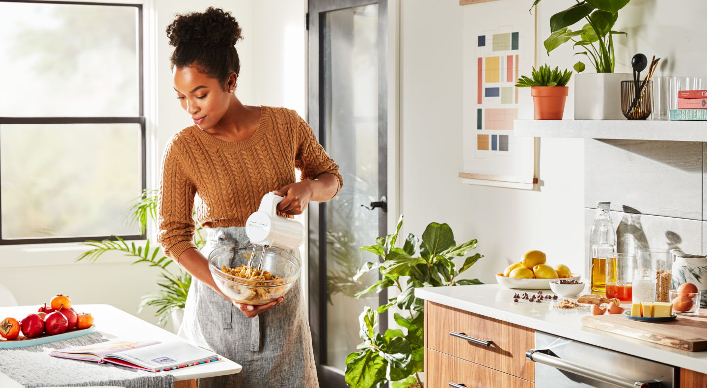 A woman in a modern kitchen reading a cookbook and holding a mixing bowl while using a Cordless KitchenAid® hand mixer to mix cookie dough. A woman in a modern kitchen reading a cookbook and holding a mixing bowl while using a Cordless KitchenAid® hand mixer to mix cookie dough.