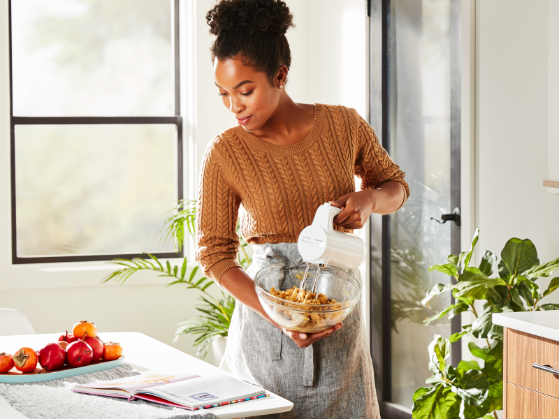 A woman in a modern kitchen reading a cookbook and holding a mixing bowl while using a Cordless KitchenAid® hand mixer to mix cookie dough. A woman in a modern kitchen reading a cookbook and holding a mixing bowl while using a Cordless KitchenAid® hand mixer to mix cookie dough.